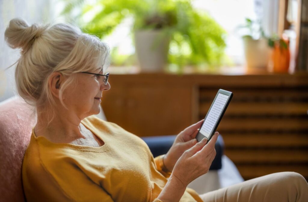 An older adult sitting on a couch, wearing reading glasses and and using an e-reader